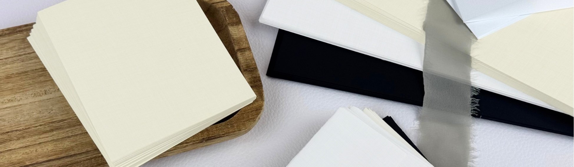 Natural White (cream) linen finish cards on a brown wooden tray next to stacked Epic Black, Ultimate White, and Natural White Linen cardstock and envelopes from LCI Paper
